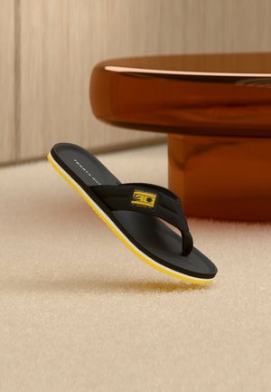 Μαύρο και κίτρινο σανδάλι σαγιονάρες με λογότυπο, τοποθετημένο υπό γωνία σε μπεζ χαλί κοντά σε στρογγυλό καφέ τραπέζι σε μινιμαλιστικό εσωτερικό χώρο.