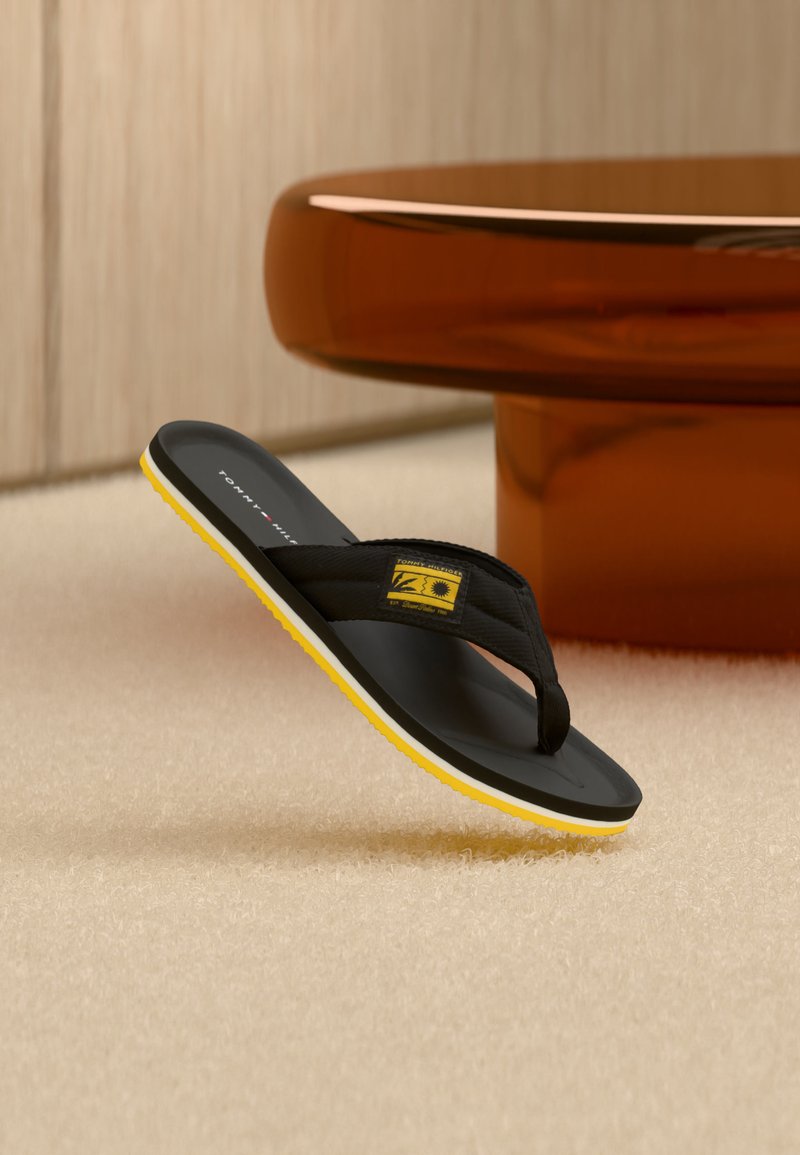 Μαύρο και κίτρινο σανδάλι σαγιονάρες με λογότυπο, τοποθετημένο υπό γωνία σε μπεζ χαλί κοντά σε στρογγυλό καφέ τραπέζι σε μινιμαλιστικό εσωτερικό χώρο.