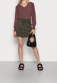 Blouse rouge foncé à motifs floraux avec un décolleté en V, associée à une jupe en velours côtelé verte avec des boutons à l'avant, et un sac à main noir tressé.