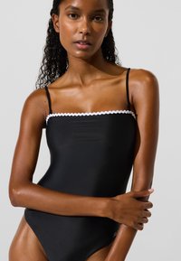 Sieviete ar cirtainiem matiem, tērpusies melnā viengabala peldkostīmā ar plānām siksnām un baltu viļņveidīgu apdari, pozē ar sakrustotām rokām.