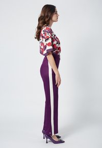 Bluzka z printem kwiatowym o szerokich rękawach, zestawiona z wysokim stanem fioletowych spodni z białymi paskami po bokach oraz dopasowanymi fioletowymi szpilkami.