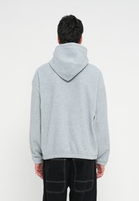 Persoană văzută din spate purtând un hanorac cu glugă de culoare gri deschis și pantaloni negri cu cusături albe, stând în fața unui fundal alb simplu.