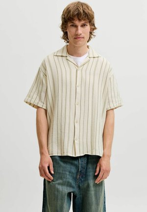 Jeune homme aux cheveux blonds ondulés porte une chemise crème à manches courtes avec des rayures verticales sur un t-shirt blanc et un jean bleu.