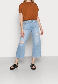 Jeans a gamba larga blu chiaro con strappi frontali, abbinati a una maglietta corta marrone e sandali neri con cinturini. Design semplice e vestibilità comoda.