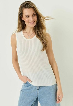 Žena s dlouhými blond vlasy, oblečená v bílém pleteném topu bez rukávů a světle modrých džínách, usmívající se a dívající se stranou.