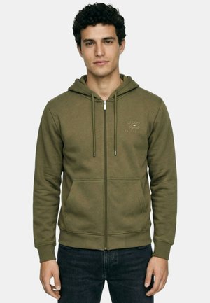 Jeune homme portant un sweat à capuche zippé vert olive avec poches avant, cordons de serrage et un texte clair sur le côté gauche de la poitrine, assorti à un jean foncé.