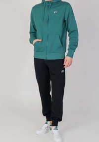 Teal zip-up hoodie med dragsko och sidofickor, kombinerad med svarta sweatpants med vita detaljer och en logotyp, fullända med vita sneakers.
