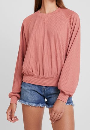 Femme portant un pull texturé rose poudré à manches longues et un short en jean bleu effiloché, debout devant un fond uni.