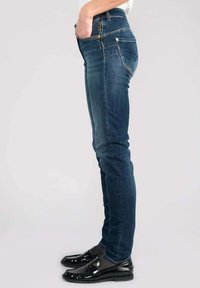 Vaqueros de mezclilla azul con un ajuste slim, cintura alta, costuras doradas y bolsillos frontales. Combinados con mocasines negros brillantes y calcetines de diseño.