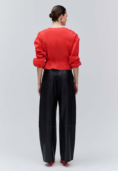 Kobieta z włosami upiętymi w kok, nosząca srebrne kolczyki, czerwoną bluzkę z długim rękawem i bufkami, czarne spodnie o szerokich nogawkach oraz czerwone szpilki, odwrócona tyłem.