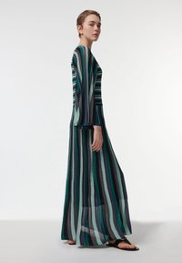 Robe longue à rayures avec un tissu transparent et fluide dans des nuances de vert, noir et gris. Dotée de manches longues et d'une coupe décontractée, portée avec des sandales noires.