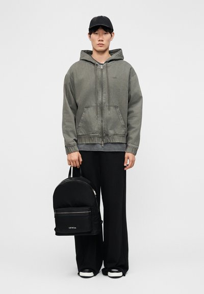 Homme portant un sweat à capuche zippé vert délavé, une casquette noire, un pantalon noir ample, des baskets blanches et noires, tenant un sac à dos noir Off-White par la poignée.