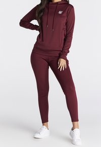 Bordeaux hættetrøje med snore og logo, parret med matchende tætsiddende leggings. Hvide sneakers fuldender det atletiske look. Glat stofstruktur.