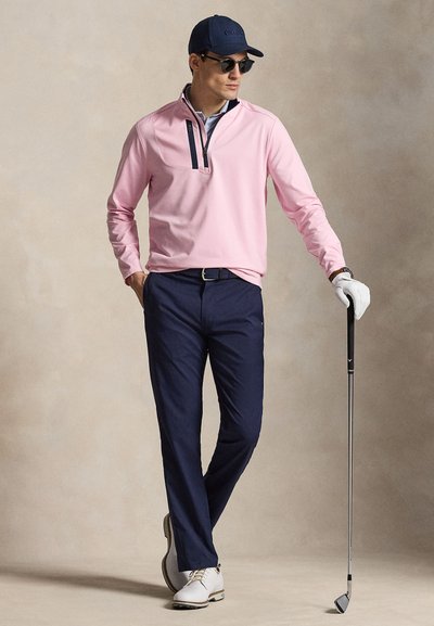 Άνδρας που φοράει ροζ πουλόβερ, μπλε σκούρο παντελόνι, λευκό γάντι γκολφ, καπέλο και γυαλιά ηλίου, κρατώντας μπαστούνι γκολφ ενώ στέκεται μπροστά σε μπεζ φόντο.