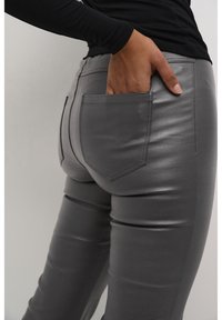 Pantalones de imitación cuero grises con un diseño ajustado, que cuentan con un bolsillo trasero y una textura suave y brillante. Estilizados con una blusa negra de manga larga.
