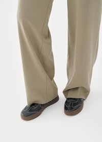 Sneakers nere con tomaia in pelle liscia, dettagli di cuciture bianche e suola in gomma marrone, abbinate a pantaloni beige larghi.