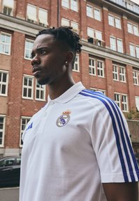 Polo bianco con strisce blu sulle spalle, con logo del Real Madrid e corona. Il tessuto sembra essere testurizzato e traspirante.