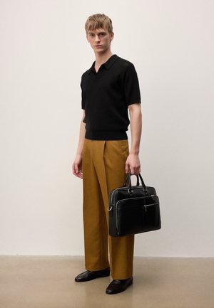 Jeune homme portant un polo en maille noire, un pantalon large moutarde, des mocassins noirs, tenant une mallette en cuir noir, debout contre un mur clair uni.