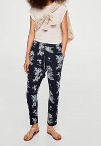 Bavlněný top v světlém béžovém odstínu, spárovaný s námořnickými kalhotami s bílým tropickým vzorem palem. Modelka má na sobě hnědé sandály a pletený šátek.