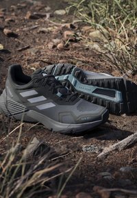 Μαύρα και γκρίζα παπούτσια τρεξίματος Adidas Terrex με ανθεκτικές εξωτερικές σόλες, τοποθετημένα σε βραχώδες, χώμα με ξερό γρασίδι.
