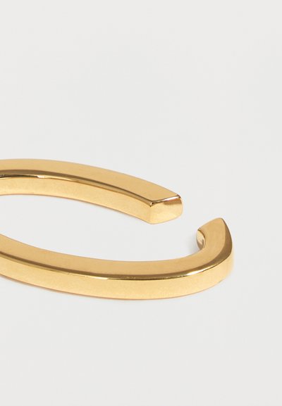 Καμπυλωτό, πολυτελές βραχιόλι από χρυσό μέταλλο με στενό άνοιγμα σε ανοιχτό φόντο.