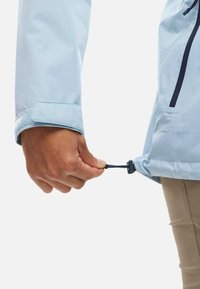 Hellblau wasserabweisende Jacke mit strukturiertem Oberflächen. Eine Hand zieht an einem verstellbaren Kordelzug am Saum, der einen schwarzen Stopper hat.