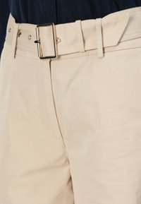 Vaahto beige -kangasvyö, jossa on suorakulmainen kultainen solki, ommeltu yksityiskohta ja viisi säätöaukkoa, kiinnitetty vaaleanvärisiin housuihin.