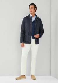 Abrigo navy con cuello, dos bolsillos frontales, llevado sobre un suéter azul. Pantalones blancos y zapatos slip-on beige completan el atuendo.