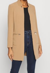 Beige dlouhý kabát s rovný střih, předními kapsami na zip a stojáčkovým límcem. Nosený přes černý top a modré džíny. Hladká textura látky.