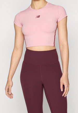 Vrouw draagt een aangesloten roze crop top met een logo en hooggetailleerde bordeauxrode legging, staand tegen een effen achtergrond.