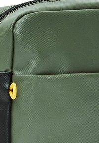 Esquina de una bolsa de nylon verde con un acabado texturizado, que presenta una sección negra y una pestaña de cremallera amarilla para un fácil acceso.