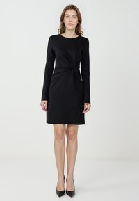 Czarna sukienka z długim rękawem o dopasowanym kroju, z detalem w postaci węzła w talii oraz gładkim wykończeniem materiału. Uzupełniona czarnymi szpilkami.