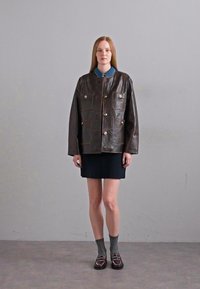 Bruine leren jas met vier voorzakken en gouden accenten, gedragen over een blauwe kraagshirt en een donkere geplooide rok, gecombineerd met grijze sokken en loafers.
