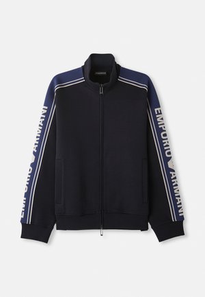 Navy blue zip-up track jacket with light beige double stripes on shoulders and sleeves, "Emporio Armani" text along both sleeves.