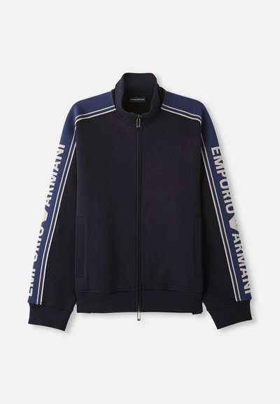 Granatowa kurtka dresowa na zamek z jasnobeżowymi podwójnymi paskami na ramionach i rękawach, z napisem "Emporio Armani" na obu rękawach.