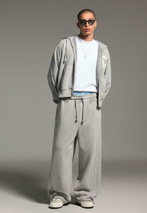 Young person wearing oversized light grey hoodie and sweatpants, white sneakers, white t-shirt, black sunglasses, and a silver necklace.