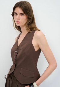 Top sin mangas marrón con escote en V, que cuenta con botones frontales, bolsillos laterales y una silueta ligeramente estructurada. Textura suave.