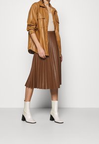 Bruine neplederen geplooide rok, beige top en crèmekleurige enkellaarsjes met zwarte hakken, gestyled met een tan knoopshirt.