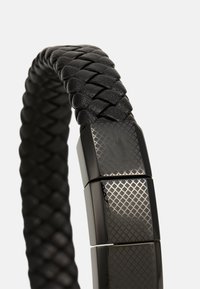 Bracelet en cuir noir avec un design tressé, présentant un fermoir en métal texturé avec une finition à motifs. Accents lisses et brillants.