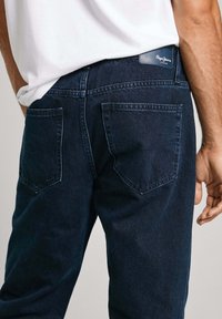 Jeans de denim azul oscuro con corte recto, dos bolsillos traseros y un parche con el logo en la cinturilla. La tela parece suave y resistente.