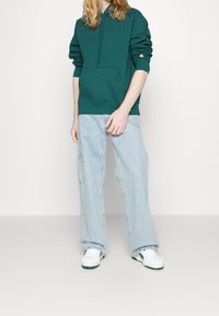 Felpa verde con tasca anteriore, vestibilità comoda. Jeans blu chiaro a gamba larga, con tasca laterale. Scarpe da ginnastica bianche con dettagli verdi.