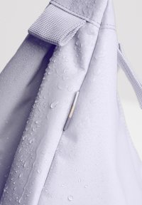 Borsa impermeabile di colore viola chiaro con superficie texturizzata, visibili gocce d'acqua e un piccolo dettaglio metallico sul lato.