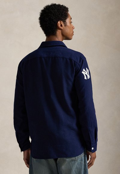 Camisa azul-marinho de tecido com mangas longas, apresentando um logo NY bordado em branco no braço direito e punhos abotoados.