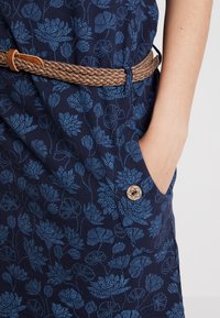 Handen är nedstoppad i fickan på en blå blommig klänning med träknappdetaljer, i kombination med ett flätat brunt bälte i midjan.