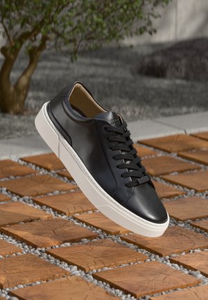 Sneaker in pelle nera con suola bianca, lacci piatti e dettaglio logo sottile, posizionata sopra una superficie lignea strutturata con sassi.