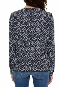 Donna che indossa una blusa a maniche lunghe blu navy con un motivo floreale bianco e azzurro chiaro, abbinata a jeans blu, vista da dietro.