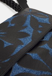 Schwarz-blaue Tasche mit einem strukturierten Lederklappen, die über einem Denim-Boden mit ausgeschnittenen Blättern-Designs versehen ist; beinhaltet einen geflochtenen Griff.
