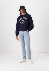 Sudadera marina con el estampado "NEW YORK GAP ATHLETIC DEPT.", jeans azul claro y zapatillas blancas. Un sombrero tipo cubo negro completa el conjunto.