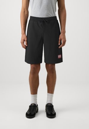 Homme portant un short noir Diesel, un t-shirt gris, des chaussettes blanches et des baskets noires Boss, debout sur un fond blanc uni.