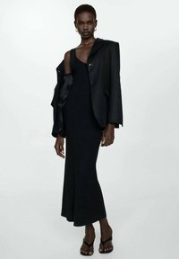 Robe midi noire à côtes associée à un blazer noir oversize avec une fermeture à un seul bouton. Le mannequin porte des sandales noires à lanières.
